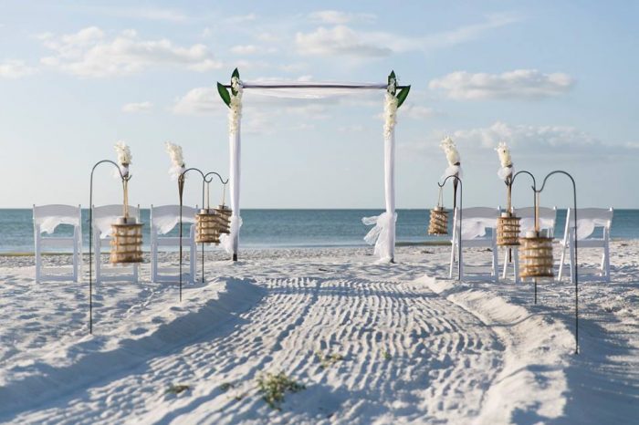 Image of a wedding arch on St. Pete beach, with three white chairs on either side and laterns hung along a brushed sand path, and the ocean in the background.