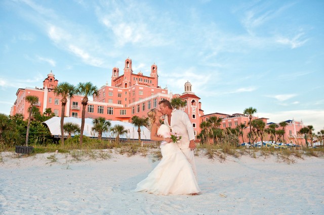 Image of a bride and groom both in white on Pass-A-Grille beach, St. Pete, with a red stone building in the background and a blue sky.